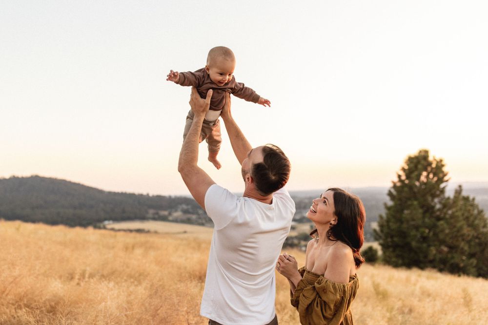 A father joyfully lifts his baby into the air while mother watches in a golden sunset meadow.