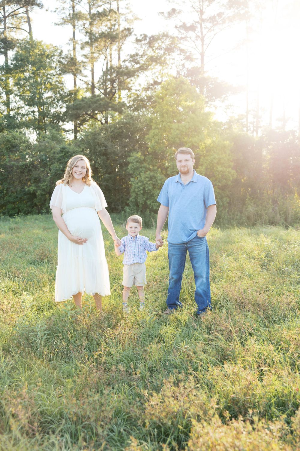 Pregnant woman in white dress and man in blue shirt hold hands with toddler boy in sunny green field.