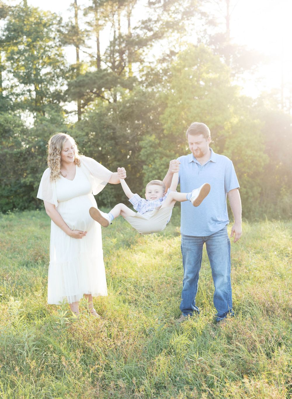 Pregnant woman in white dress and man in blue swing toddler between them in sunny green meadow.