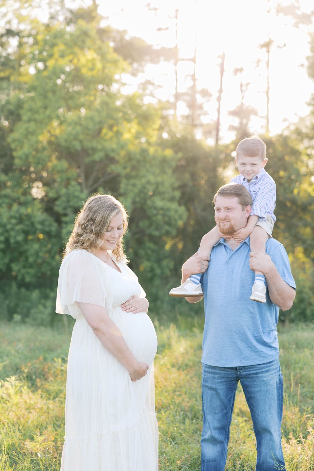 Pregnant woman in white dress with husband carrying toddler on shoulders during golden hour maternity photo session.