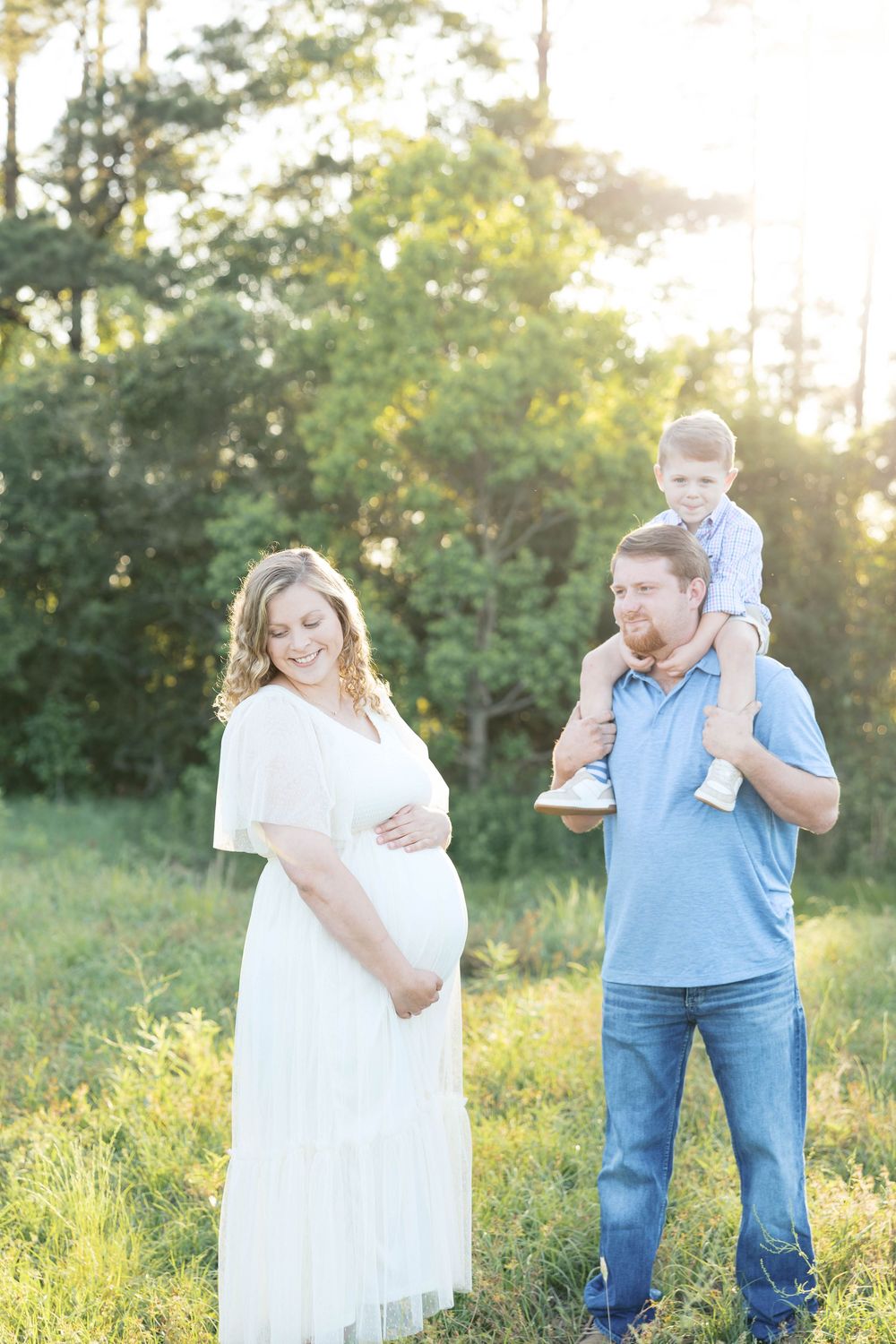 Pregnant woman in white dress smiles beside husband holding toddler on shoulders in sunny green field.
