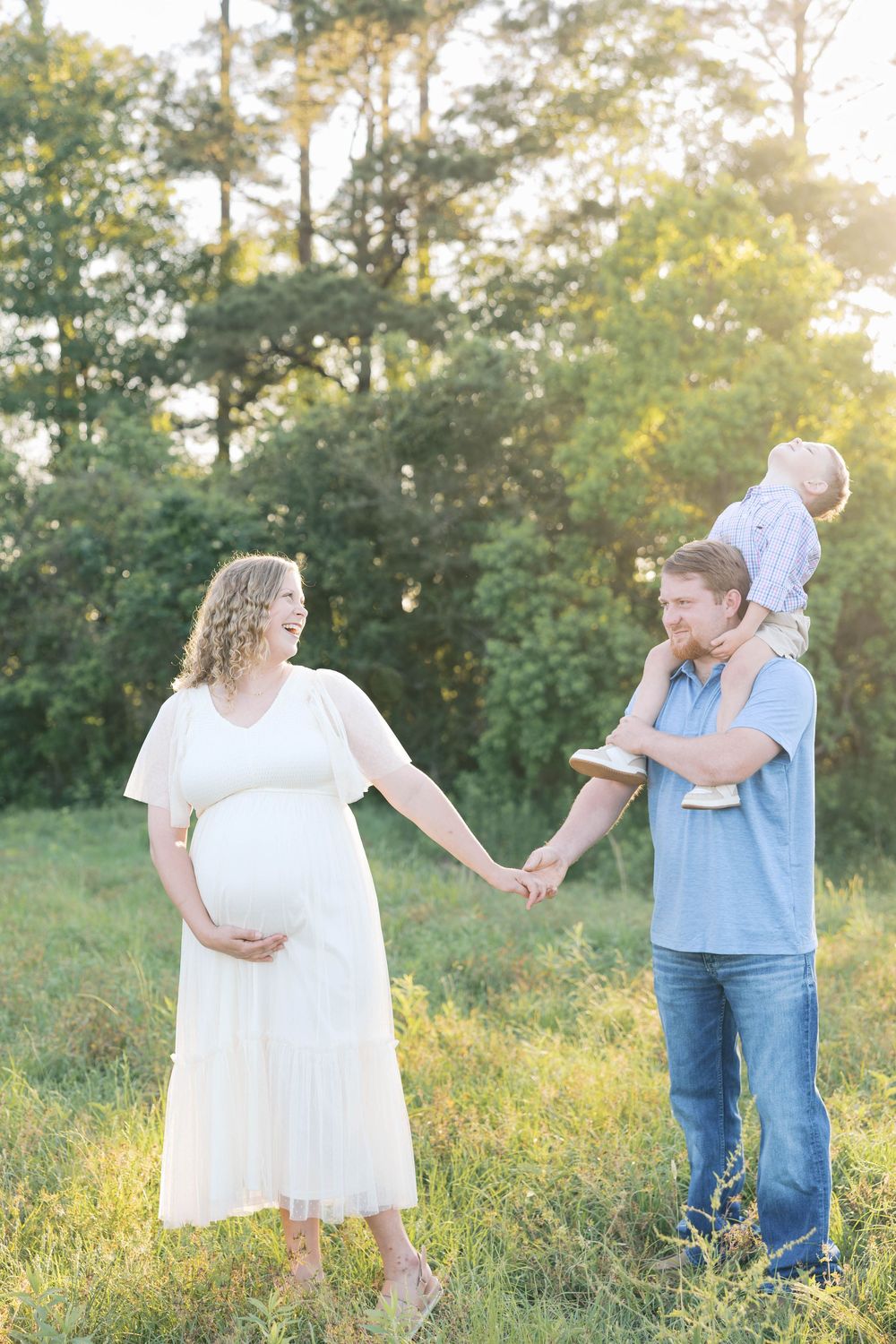 Pregnant woman in white dress holds hands with man carrying toddler on shoulders in sunny outdoor field.