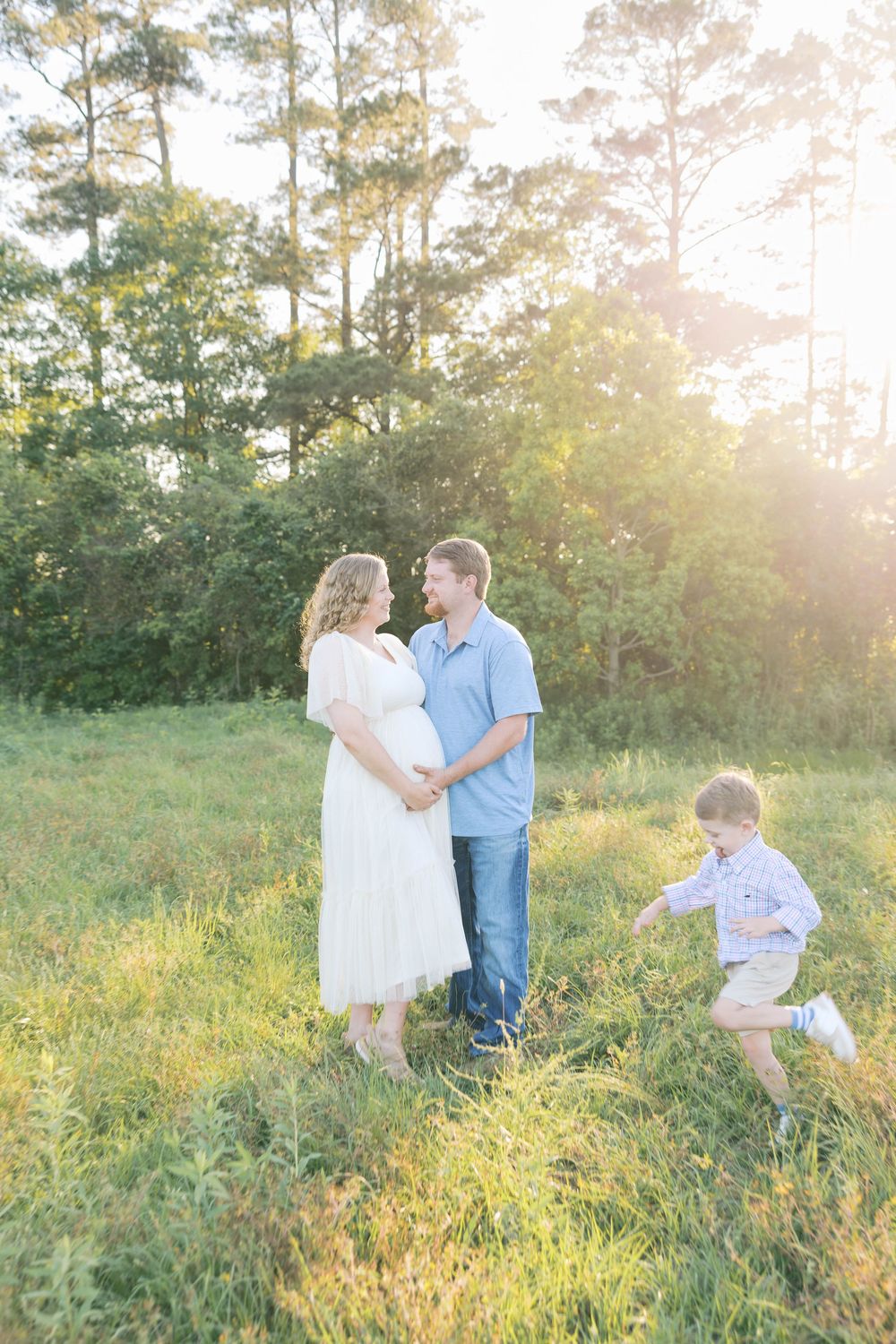 Expectant couple smiles at each other while young son plays beside them in a sunlit outdoor field.