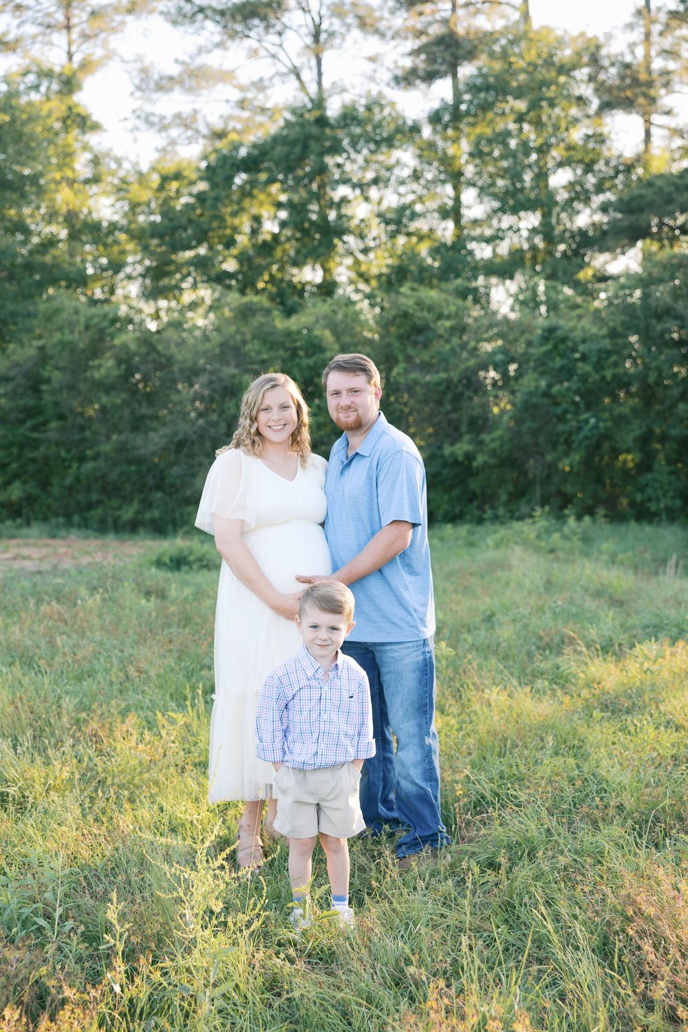Smiling pregnant woman in white dress poses with husband and young son in a lush green meadow.