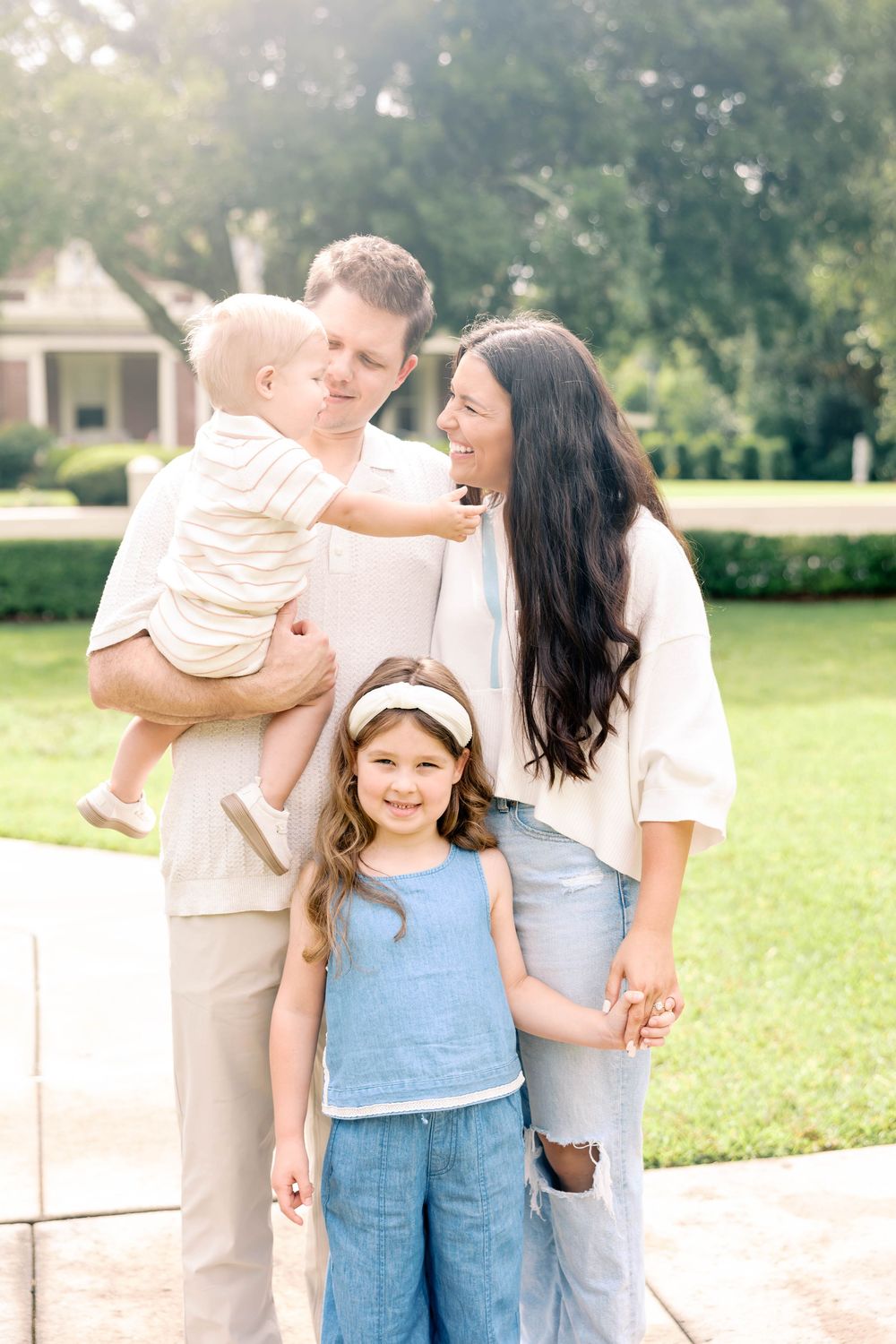 A happy family of four poses outdoors, with parents holding a baby and a young girl smiling in front.