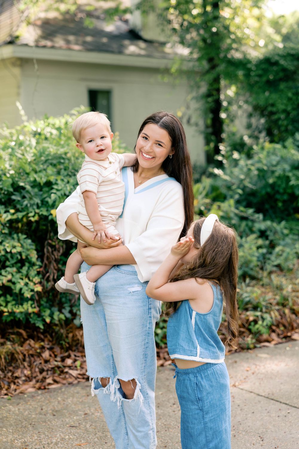 A smiling brunette mother in jeans holds a baby while her young daughter hugs her outdoors near lush greenery.