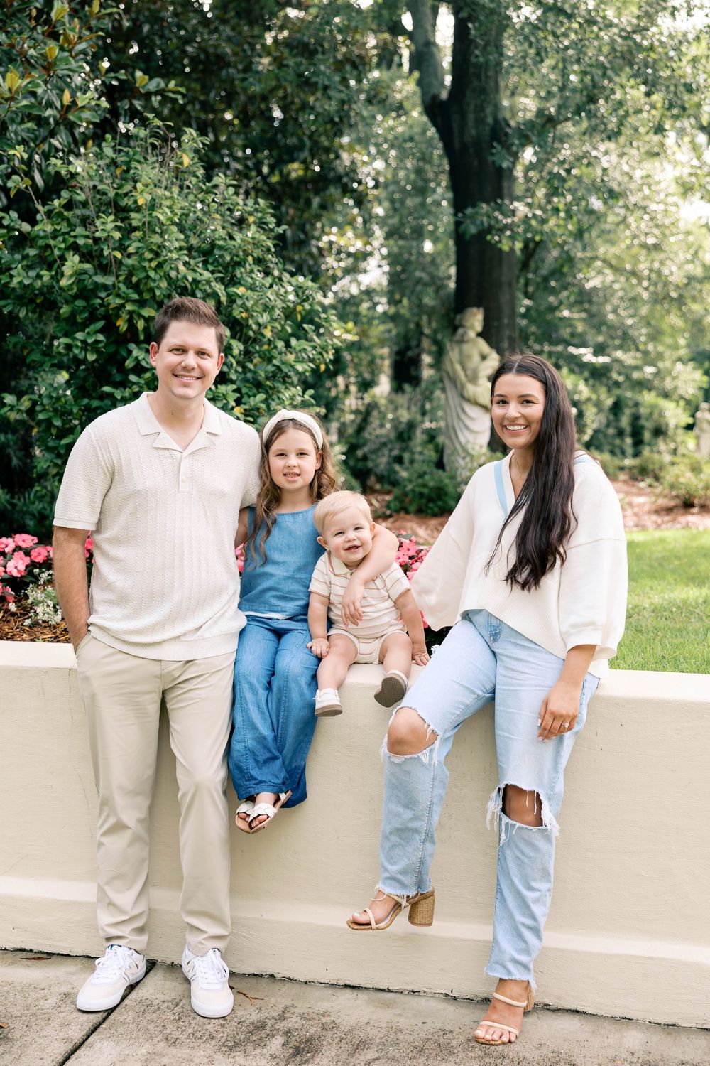 A smiling family of four poses outdoors near a garden wall, featuring two adults and two young children in casual attire.