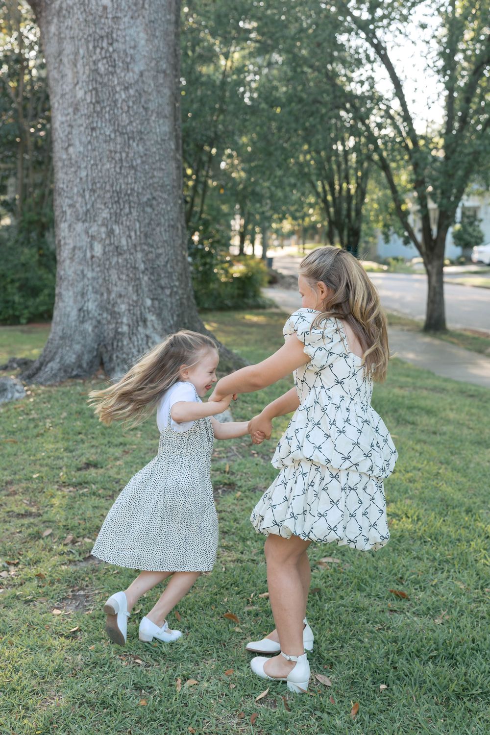 Two young girls in matching white dresses hold hands and spin together on a green lawn surrounded by tall trees.
