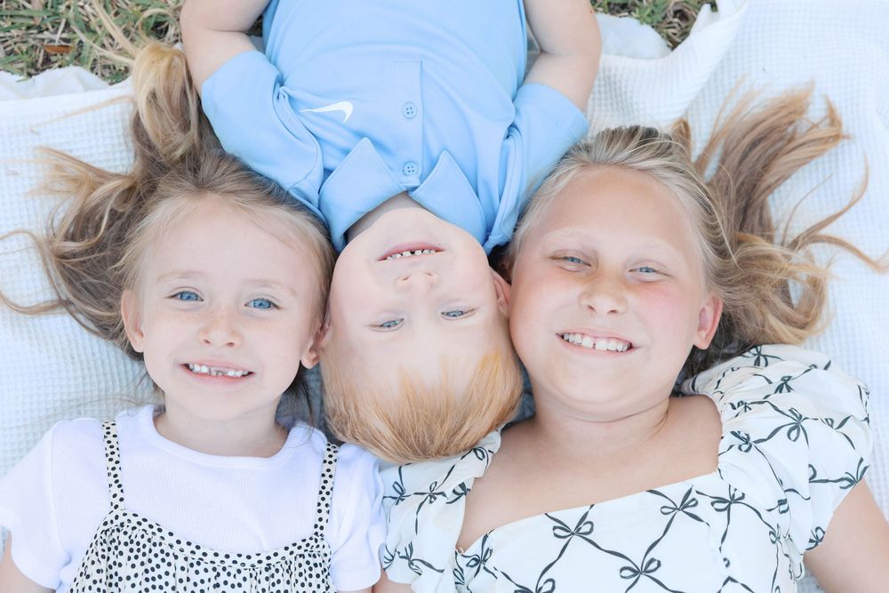 Three smiling children lying on a blanket outdoors, two girls and a boy with blonde hair enjoying a sunny day.