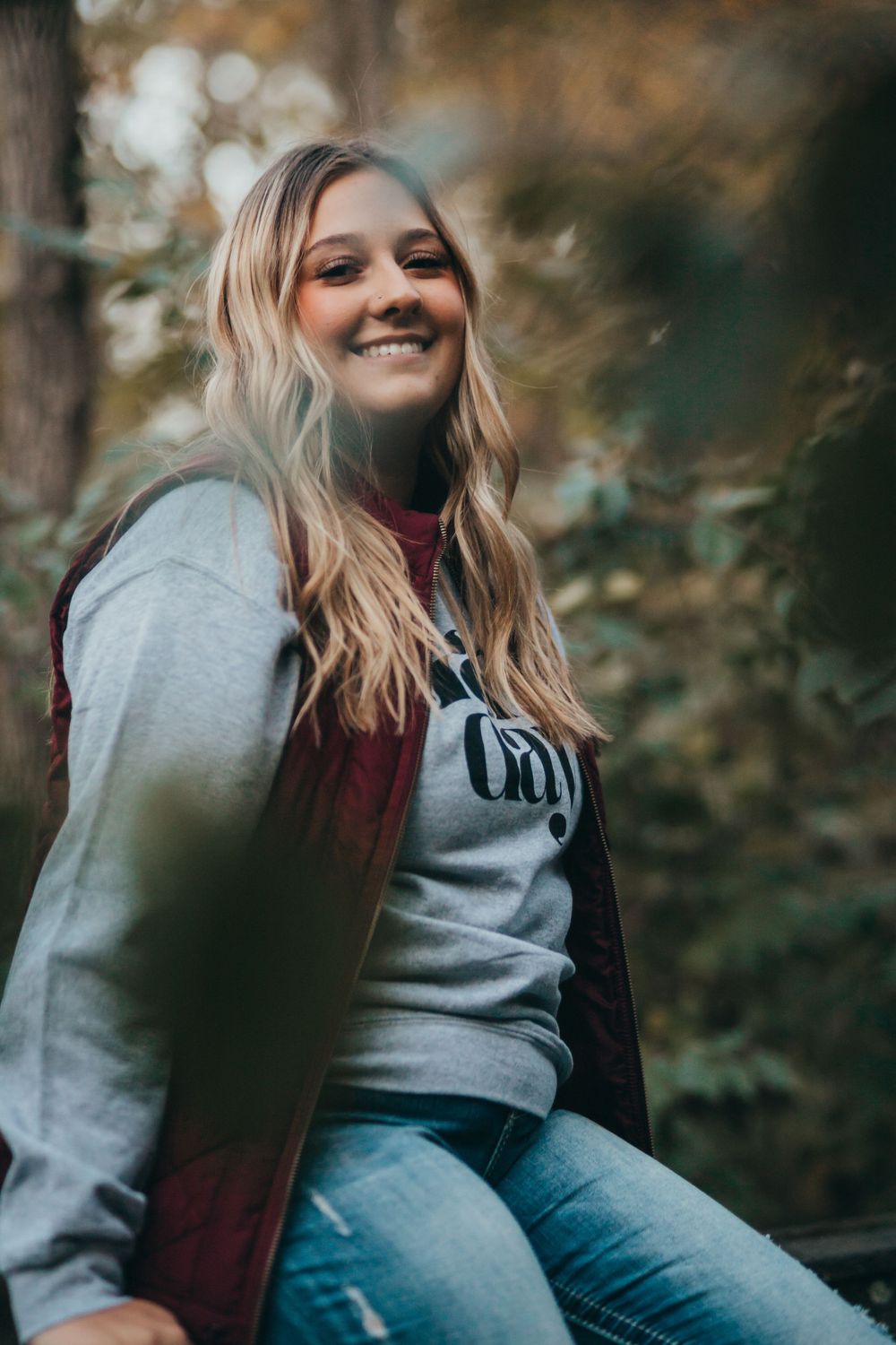 A candid outdoor portrait shows someone wearing a gray sweatshirt and burgundy vest sitting in a wooded setting.