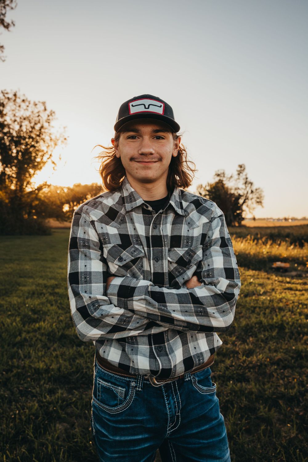 Portrait in natural lighting at sunset showing person wearing plaid flannel shirt, baseball cap and jeans in outdoor setting.