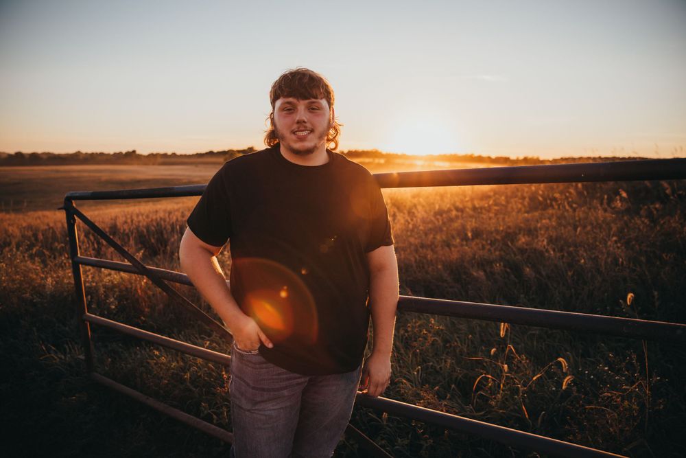 Person in black t-shirt smiling while leaning on fence railing during golden sunset over rural field.