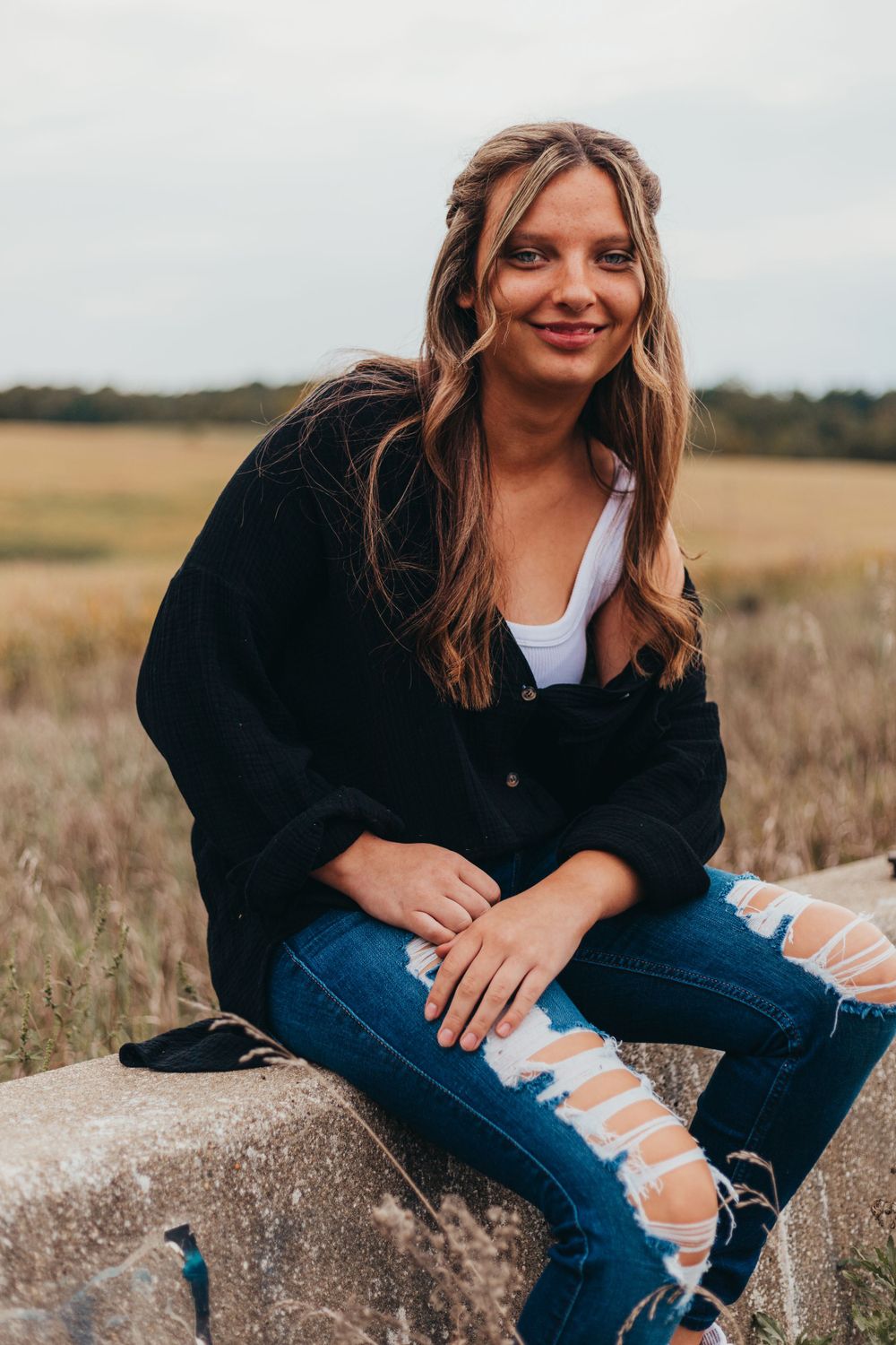 Casual autumn portrait of someone wearing distressed jeans and black cardigan sitting on rock in golden field.