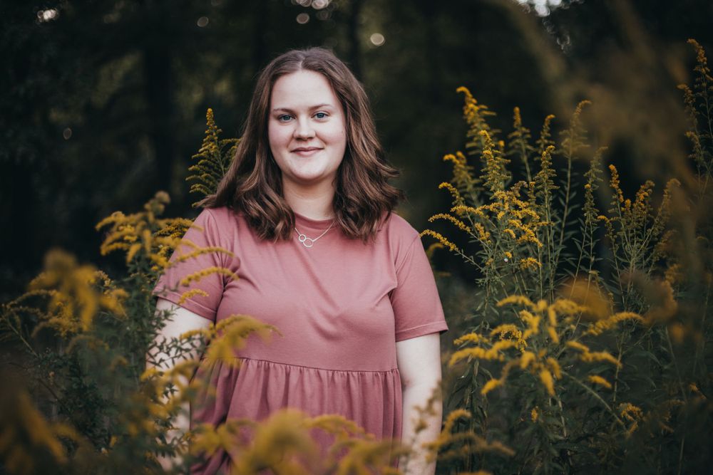 A person in a pink dress poses outdoors among tall yellow goldenrod flowers in a natural setting.