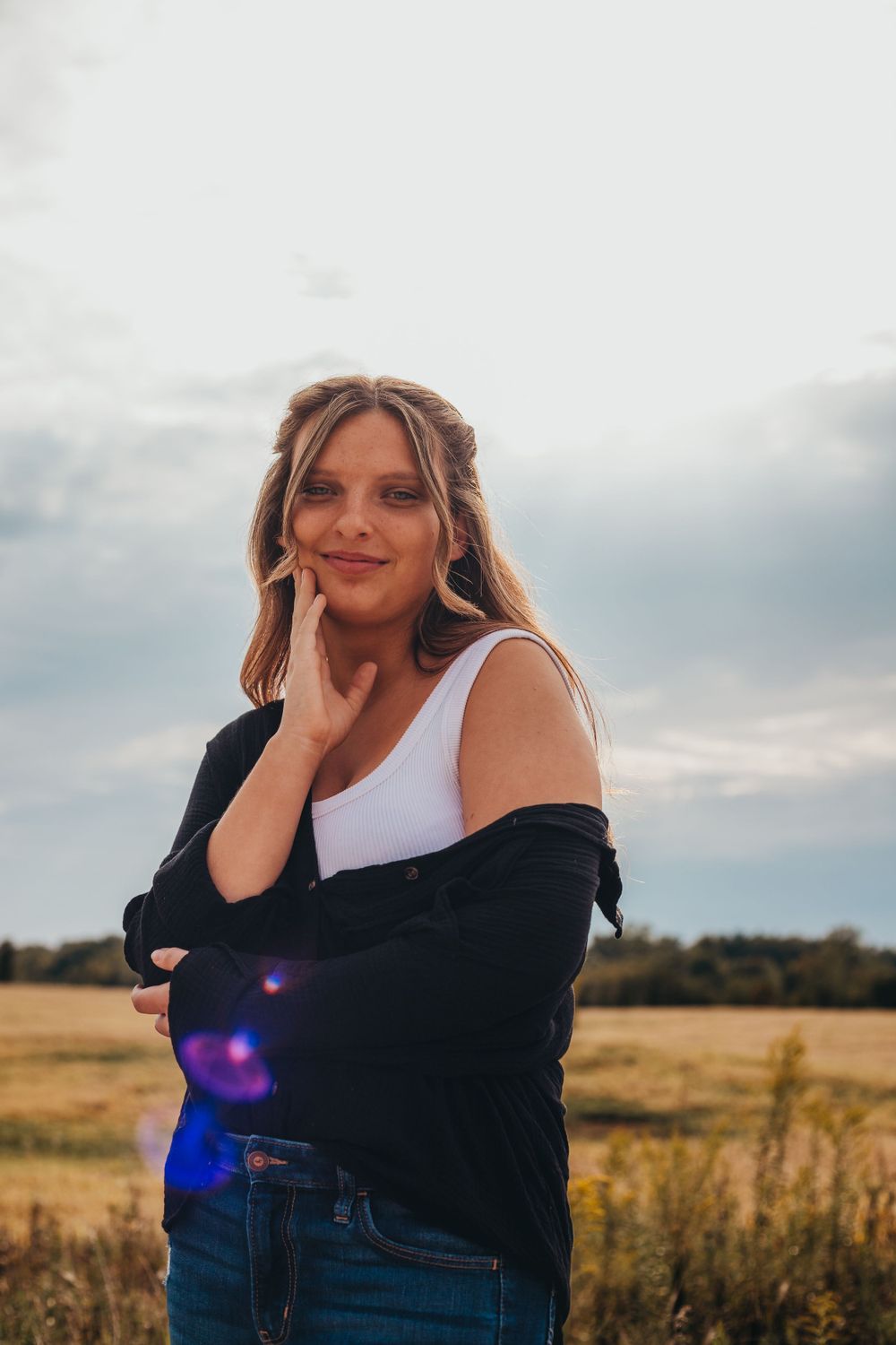 Young woman in white tank top and black cardigan poses in golden meadow at sunset with clouds in background.
