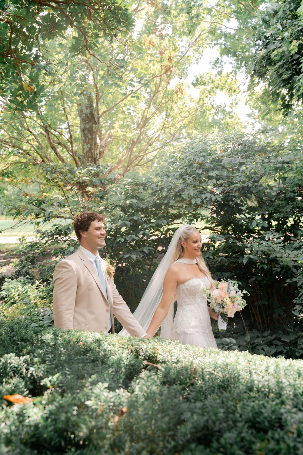 Bride and groom hold hands in a lush garden, she wears a strapless gown with veil, he wears a tan suit.