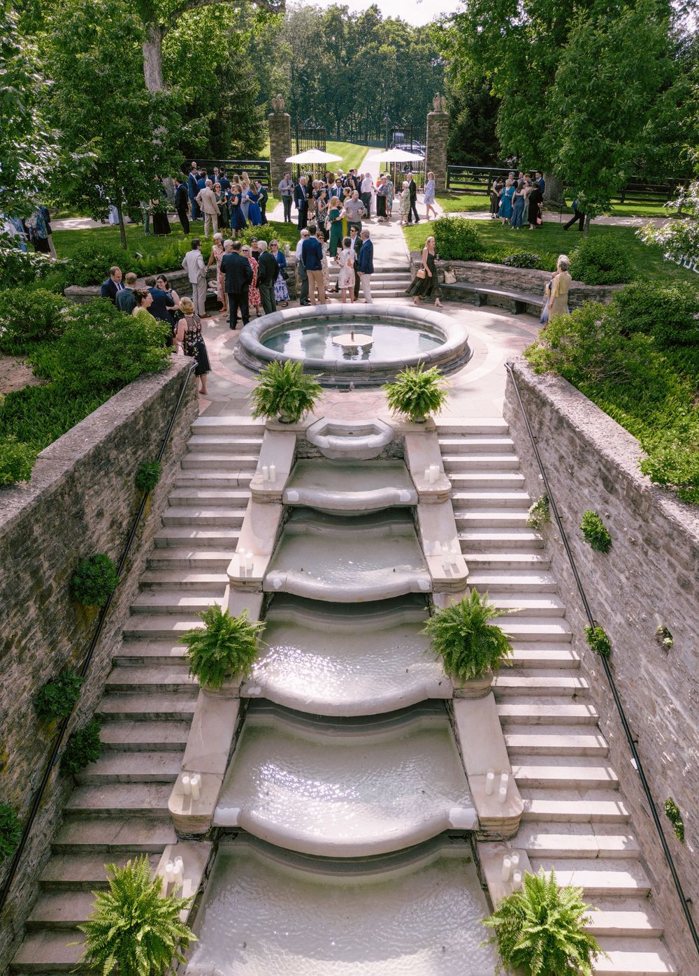 Elegant outdoor wedding reception beside a cascading stone fountain with terraced water features, lush greenery, and guests mingling.