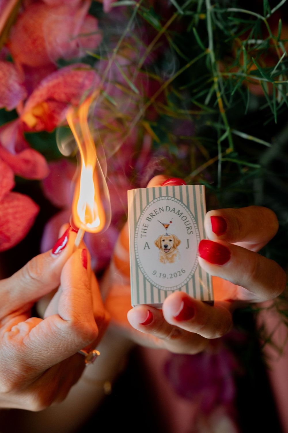 Hands with red nails lighting a custom matchbox labeled The Brendamours with a golden retriever illustration at a wedding.