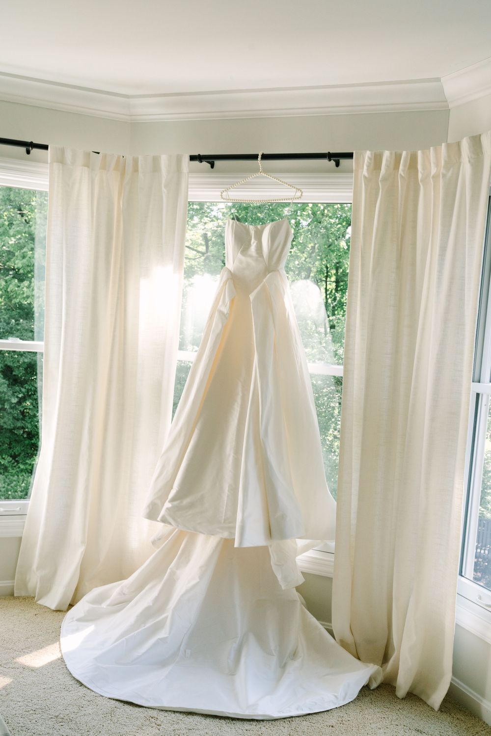 Elegant strapless white wedding gown hanging in a sunlit bay window with sheer ivory curtains.