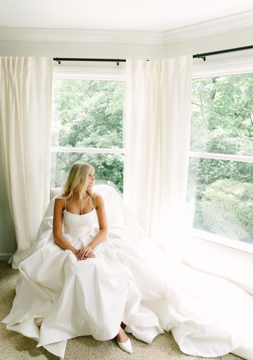 Bride in white gown sits gracefully by bay windows with sheer curtains, gazing outside at lush green trees.