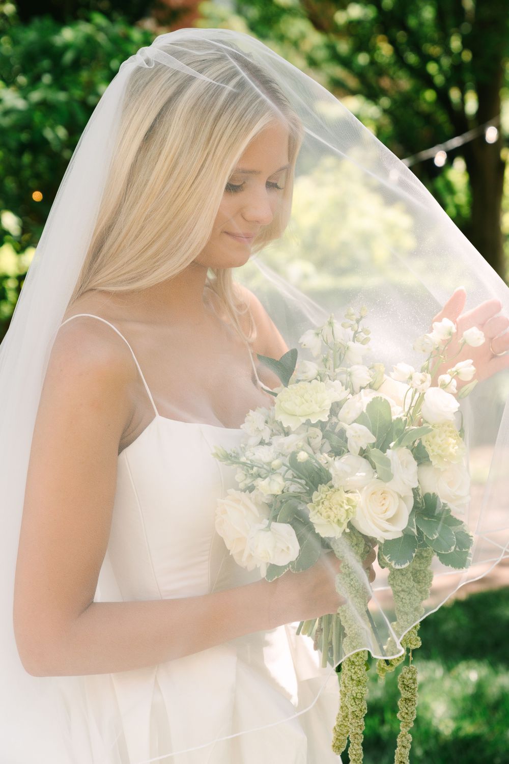 Blonde bride in white gown and veil gazes down at elegant white floral bouquet outdoors.