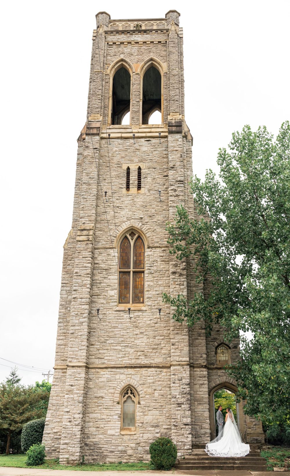 Bride and groom pose beneath a tall Gothic stone church bell tower surrounded by lush green trees on their wedding day.