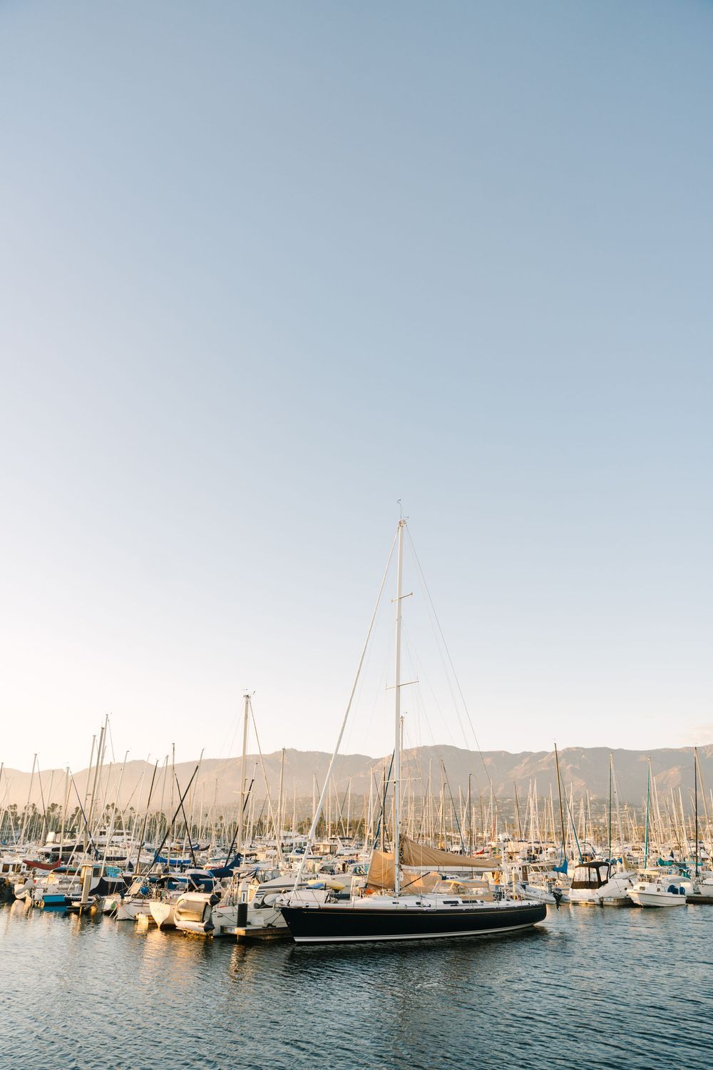 Panoramic view of sailboats and yachts docked in a marina with mountains in the background on a clear sunny day.