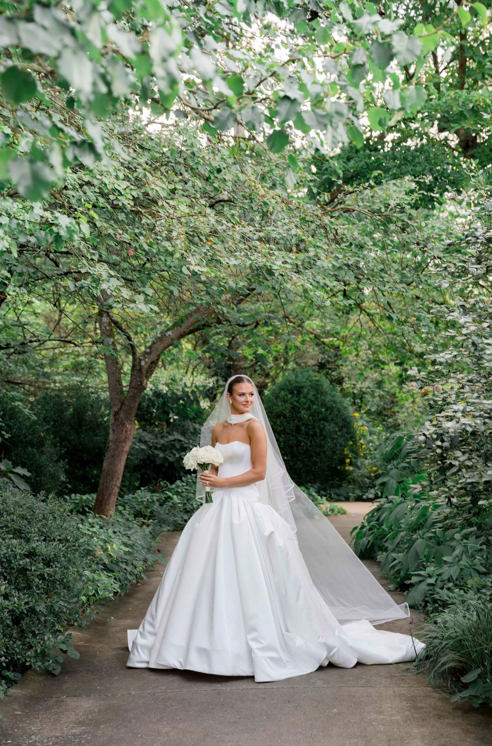 A bride in a luxurious white ball gown poses elegantly under blooming trees in a garden setting.