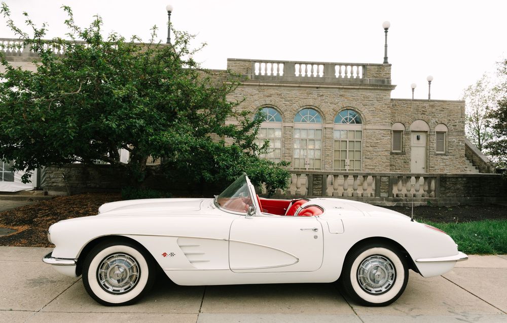 White 1959 Chevrolet Corvette convertible with red interior and whitewall tires parked in front of a stone building.