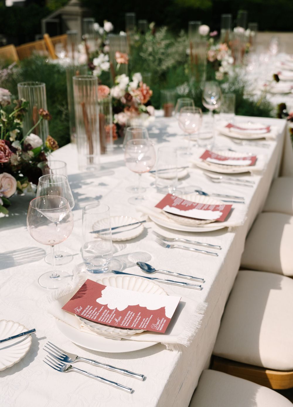 Elegant outdoor wedding reception table with white linen, burgundy menus, crystal glassware, and floral centerpieces.