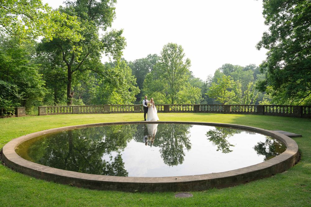 A curved stone balustrade overlooks a tranquil pond surrounded by lush green trees in a peaceful garden setting.
