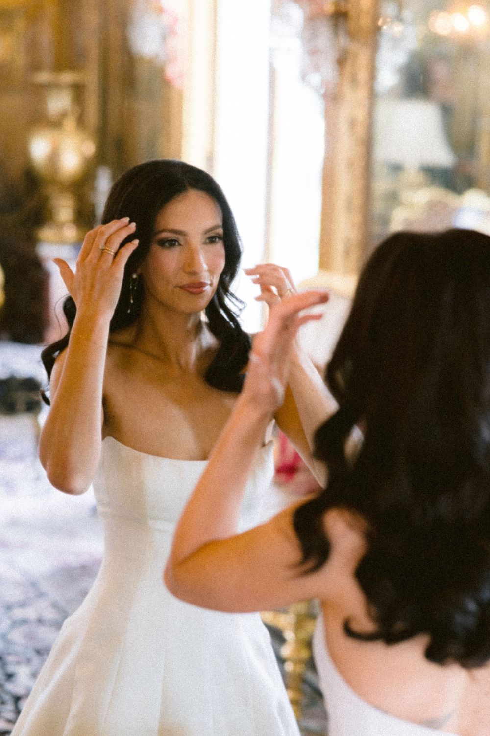 Bride in white strapless gown adjusting her hair while looking in an ornate mirror in an elegant venue.