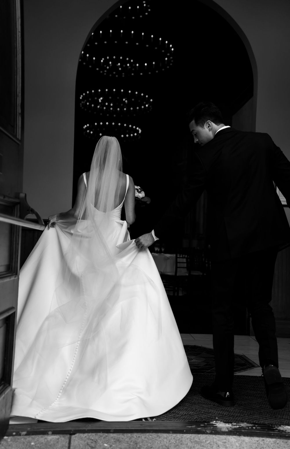 Black and white wedding photograph showing a bride in a flowing white gown and groom ascending stairs under decorative lights.