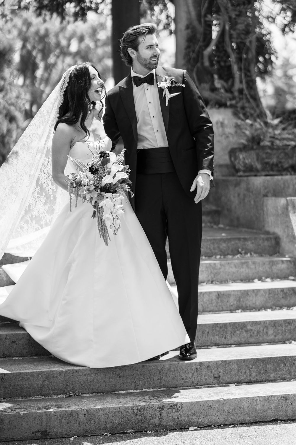 Black and white photo of a bride and groom walking down stone steps, bride holding a bouquet in a ballgown.