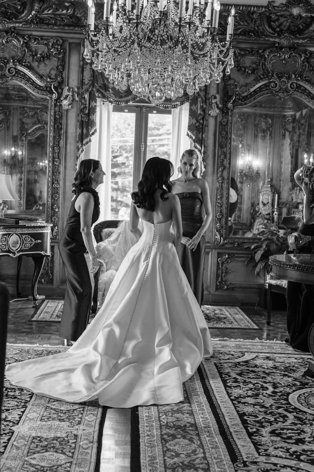 Black and white photo of a bride in a ballgown with two bridesmaids in an ornate chandelier-lit room.