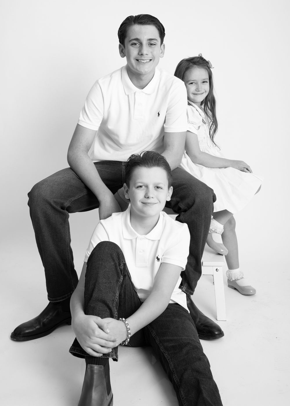 Black and white studio portrait of three people in white polo shirts sitting and posing together against light background.