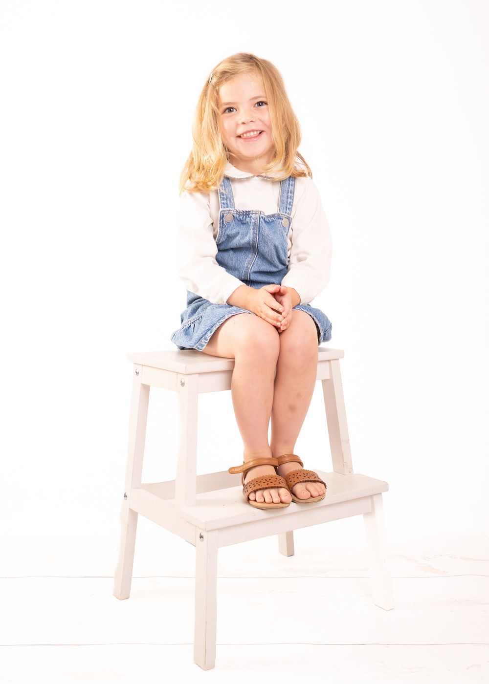 Young blonde girl in denim overalls and white shirt sits smiling on a white wooden step stool.