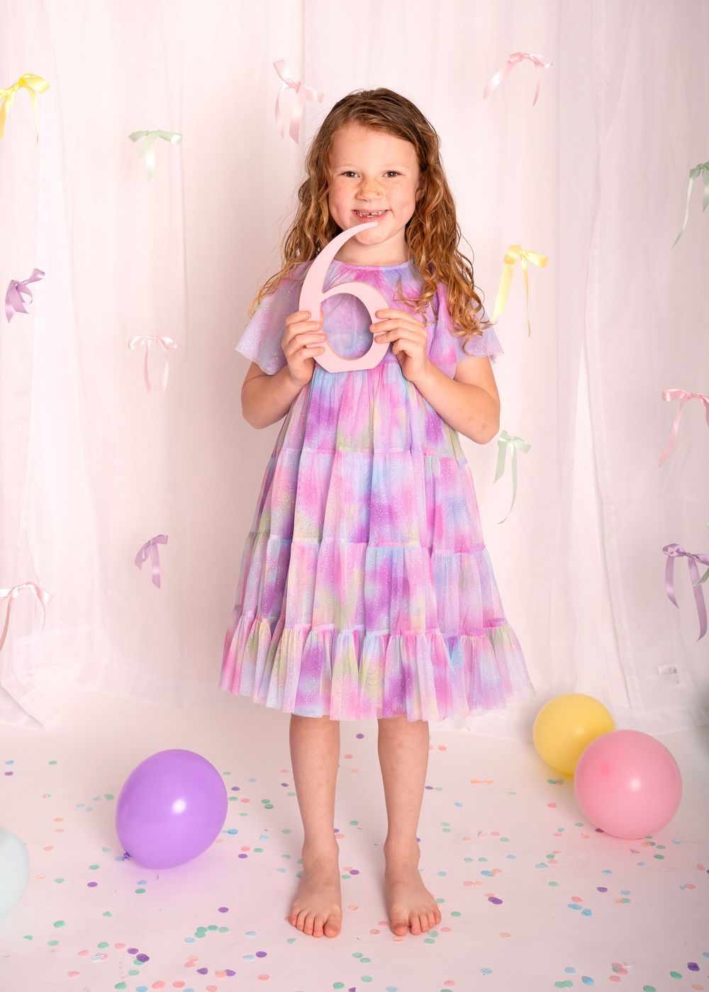 A young girl in a colorful tiered dress holds a number 6, surrounded by pastel balloons and confetti at a birthday party.