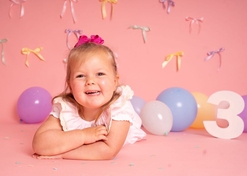 A smiling toddler girl poses with colorful balloons and a number 3 on a pink birthday photo backdrop.