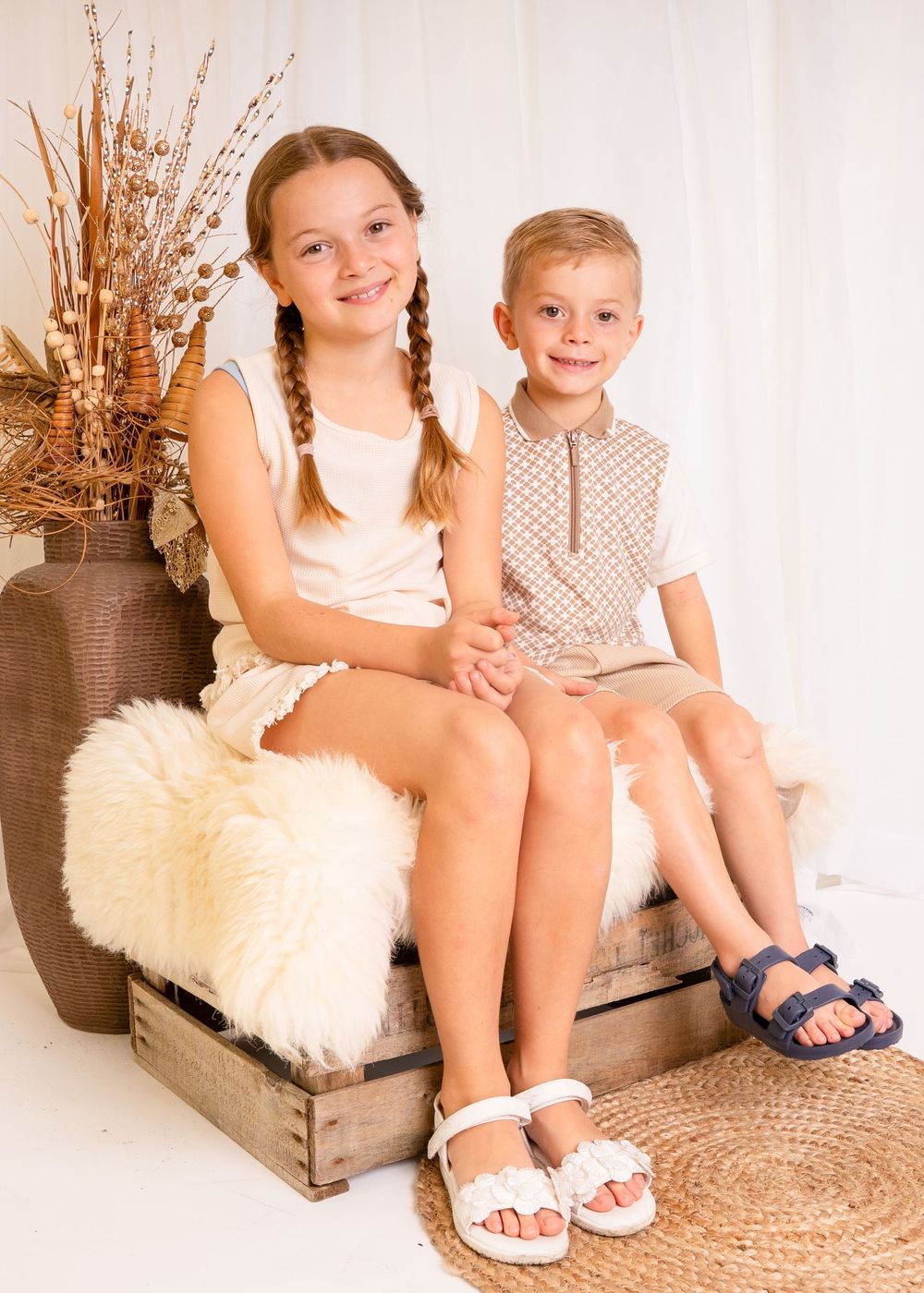 Two children, a girl with braids and a boy, sit together on a wooden crate in a bright, cozy studio setting.