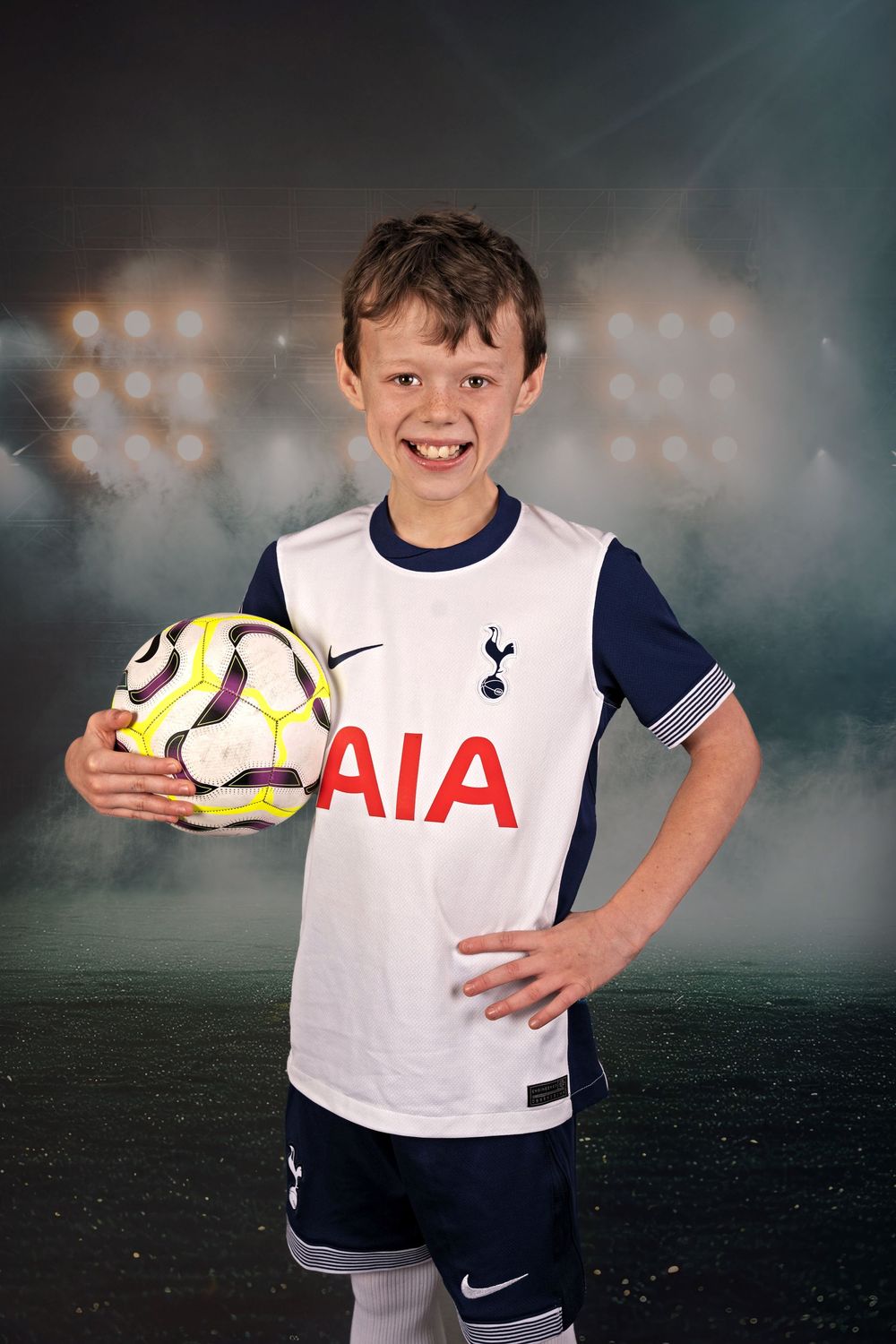 Young soccer player in white Tottenham Hotspur jersey holds ball against dark background with stadium lights.