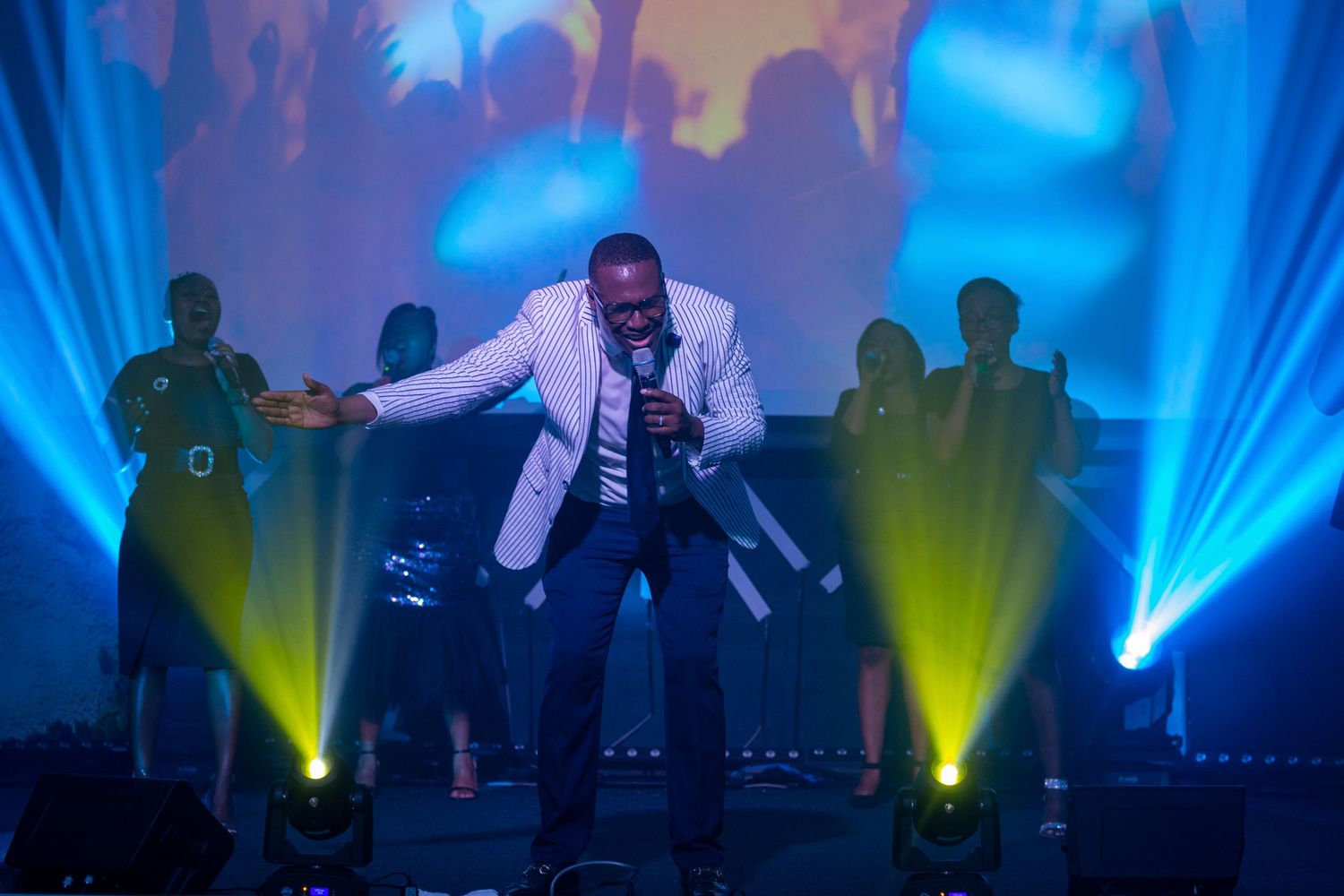 Performer in white jacket singing on stage with dramatic blue and yellow lighting effects.