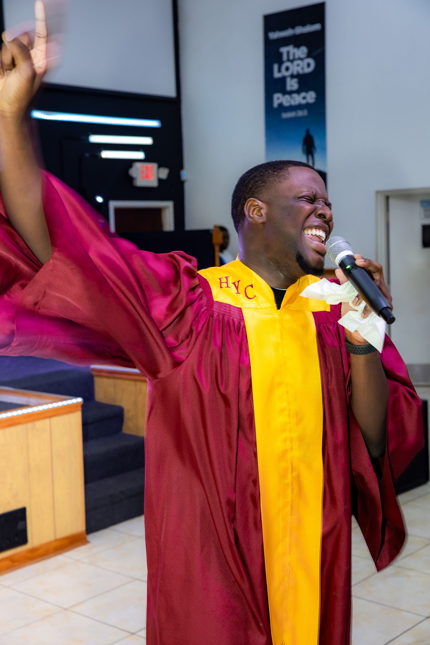 A gospel singer in maroon and gold choir robes passionately performs with upraised arms in a church setting.