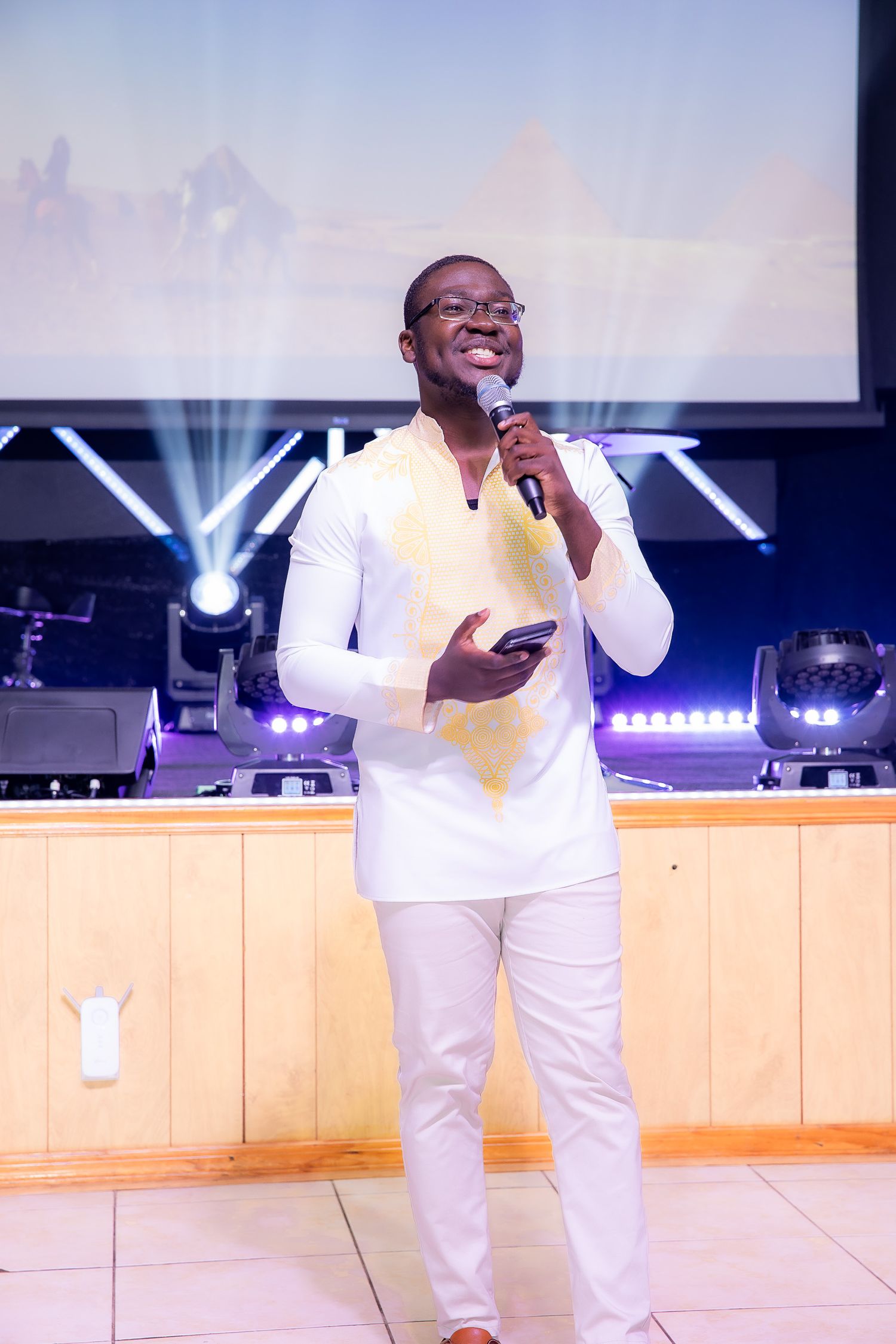 A worship leader in all white attire performs with a microphone on a church stage with lighting effects.