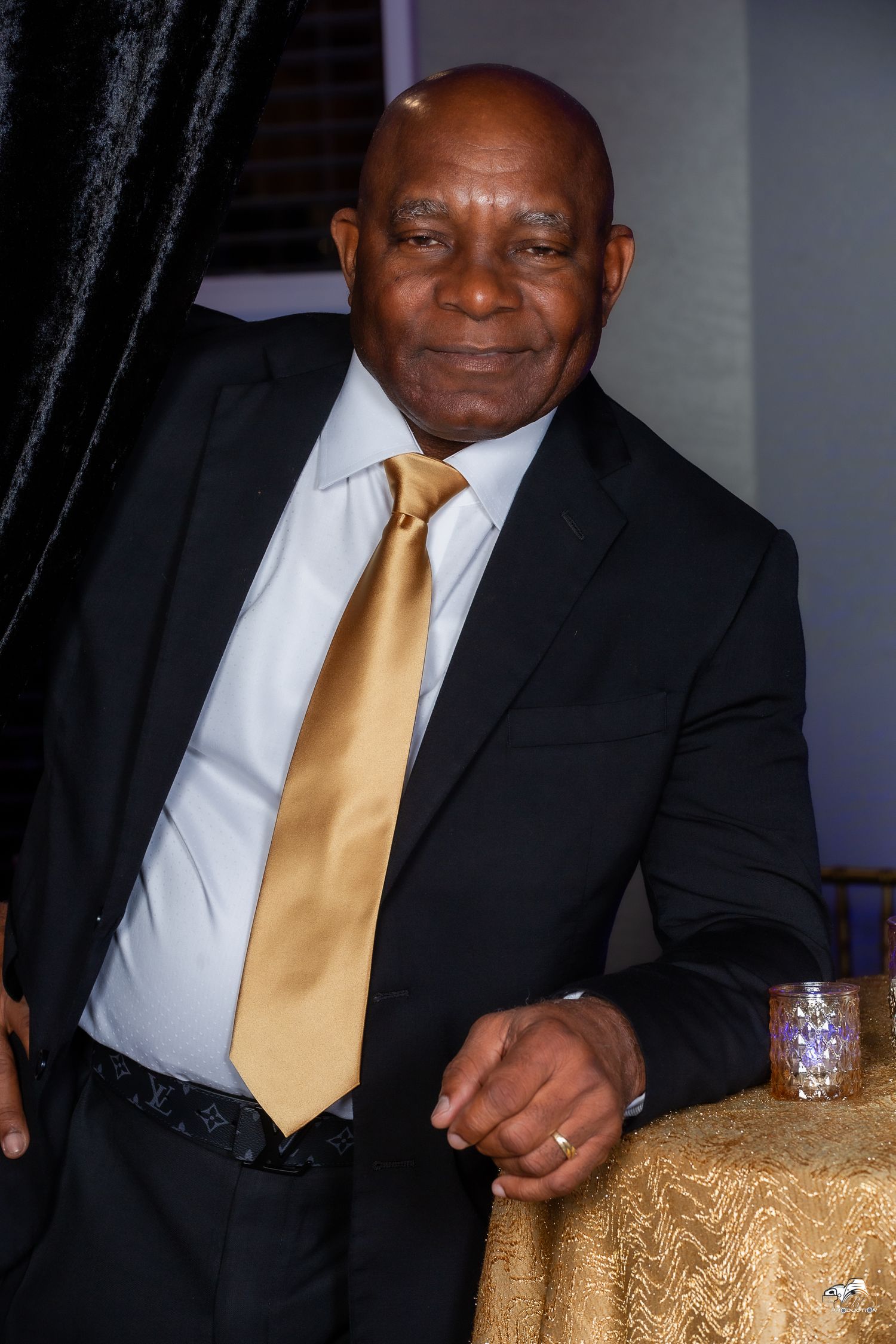 A person in a formal black suit with a gold tie smiles warmly at a celebration event.