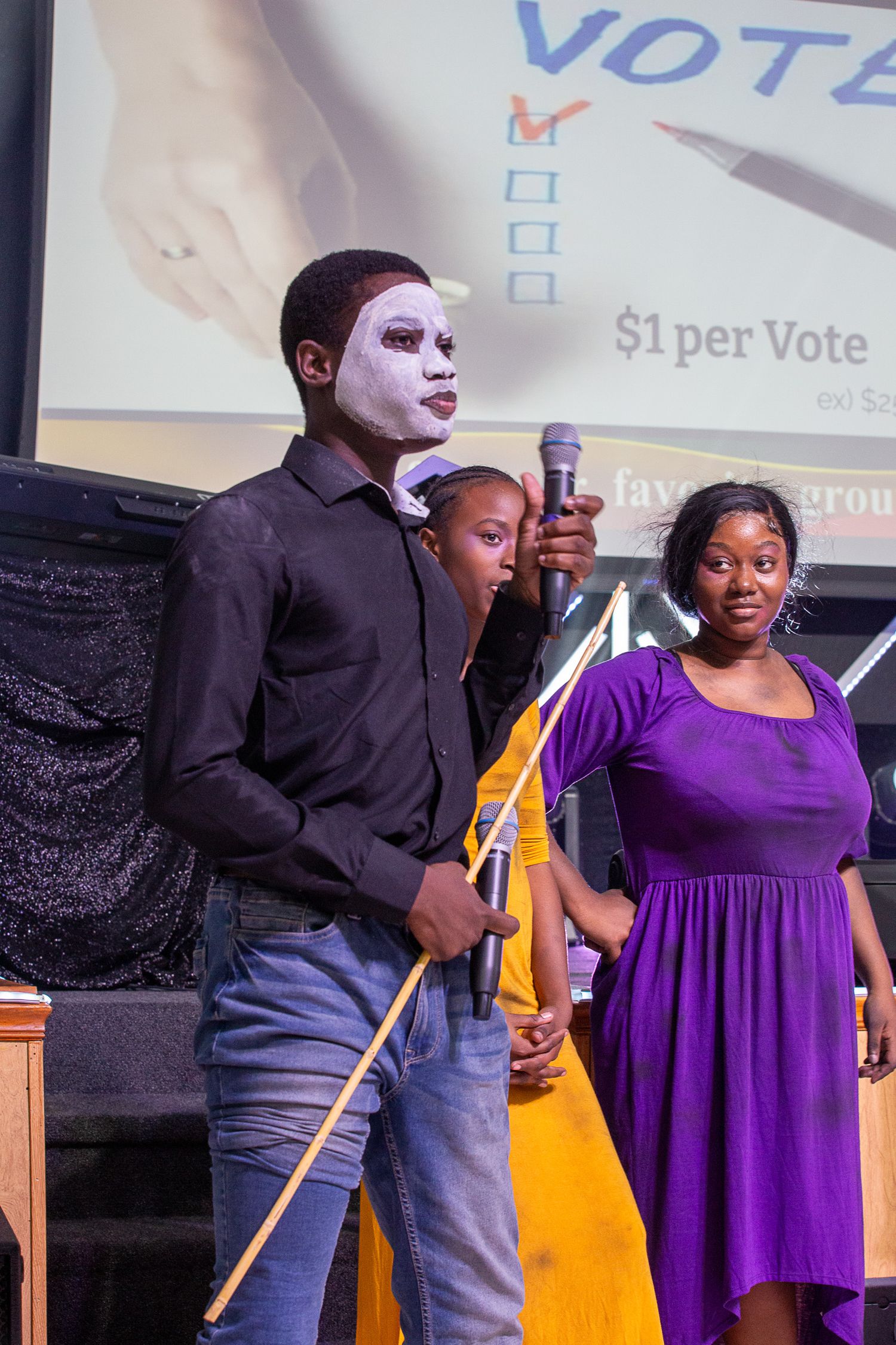 Performers in theatrical costumes stand on stage with voting information displayed on a screen behind them.