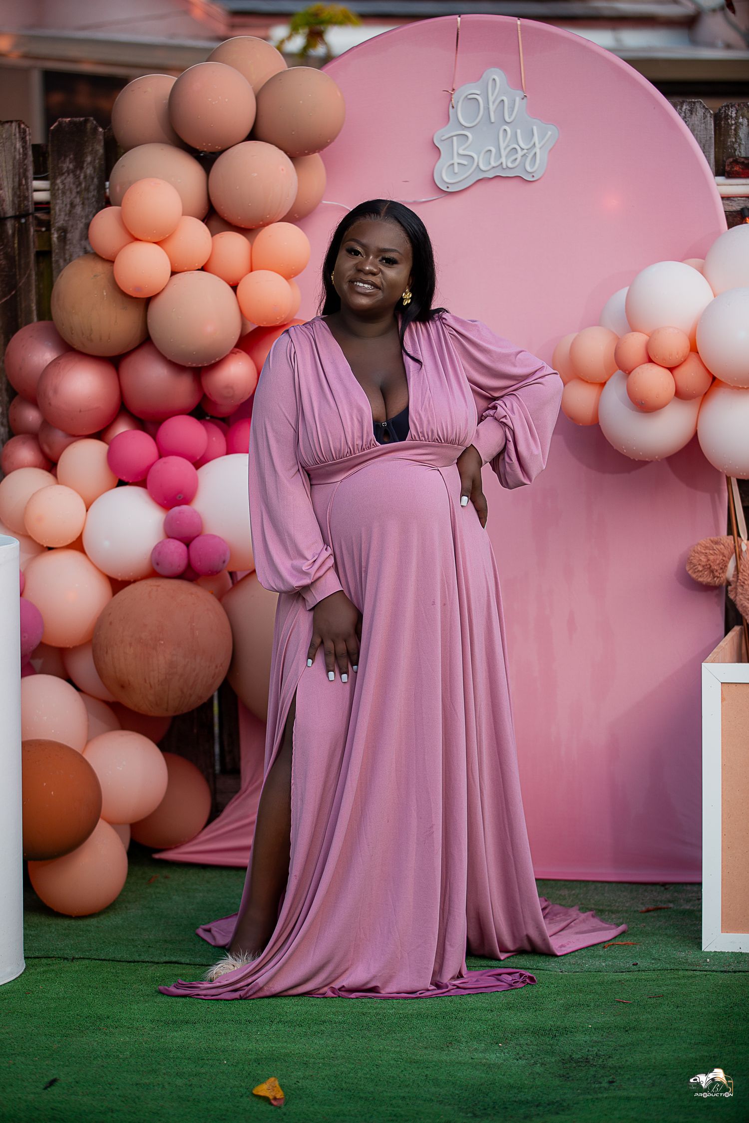 Maternity photoshoot with pink gown against balloon arch and 'Oh Baby' sign backdrop.