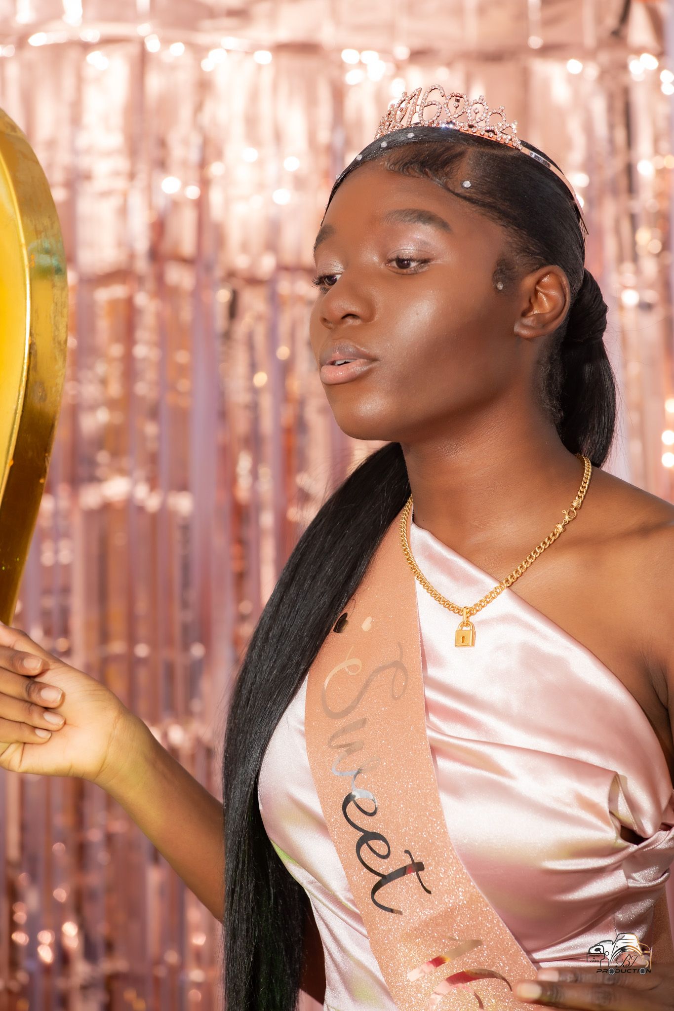 A person in a pink satin dress wears a tiara and gold necklace against a sparkly rose gold backdrop.