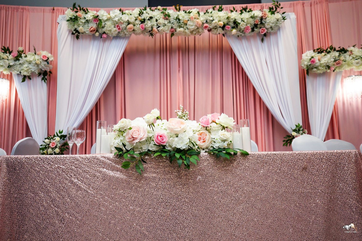 Wedding backdrop with white drapes, pink lighting and floral arrangements on a sequined table.