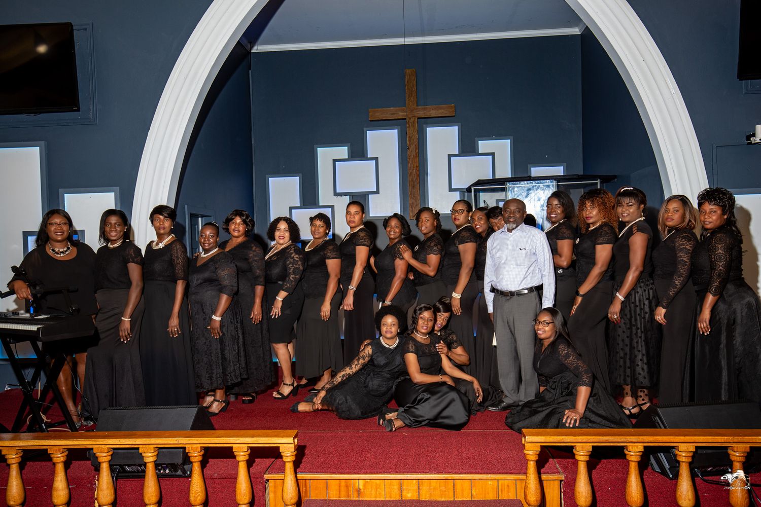 A large choir in black attire poses on stage under an arched church backdrop with a cross.